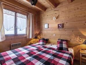 Un dormitorio de cabaña de madera con colcha a cuadros, dos almohadas, una mesita de noche, una lámpara y una ventana con cortinas.