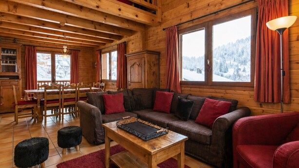 A cozy cabin interior with a wooden dining area, a sofa with red cushions, and a view of snowy mountains through large windows. - Cozy chalet for 10, fireplace, garage, WIFI, pets welcome (La Clusaz)