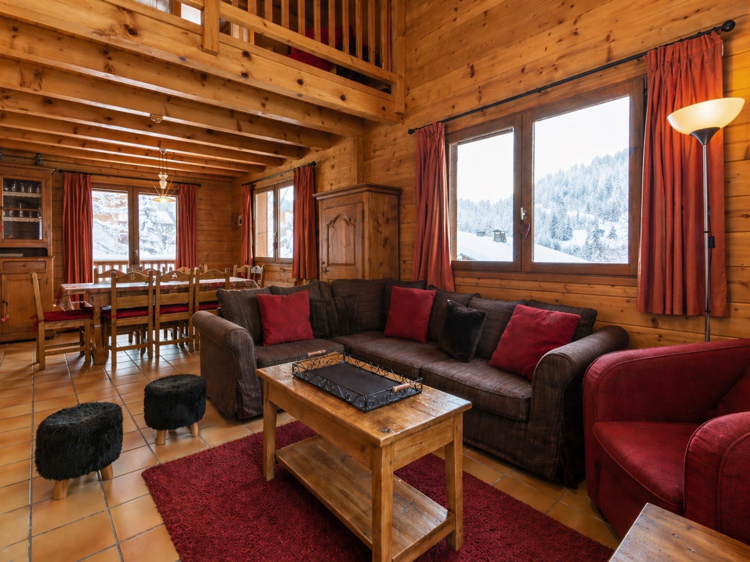A cozy cabin interior with a wooden dining area, a sofa with red cushions, and a view of snowy mountains through large windows.