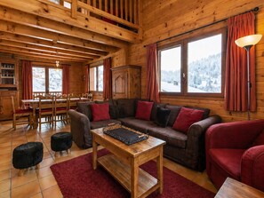 Interior acogedor de una cabaña con zona de comedor de madera, un sofá con cojines rojos y vistas a montañas nevadas a través de amplias ventanas.