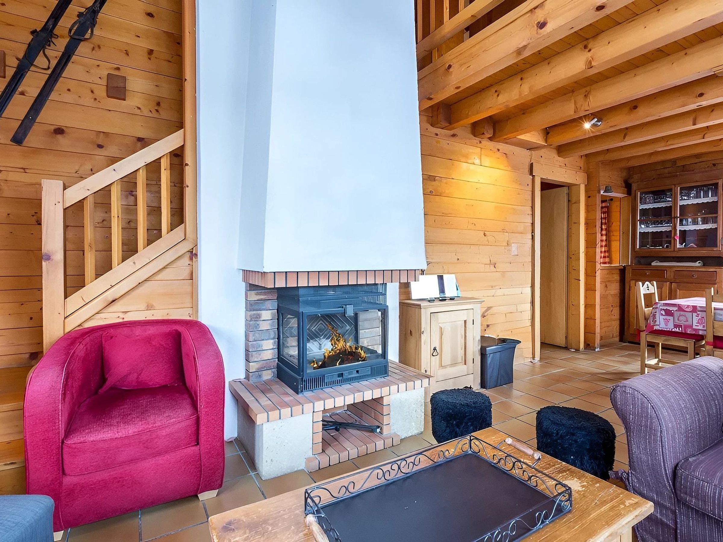 Interior de uma cabana de madeira com lareira, uma cadeira vermelha, uma mesa e uma escada.