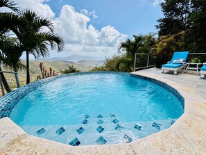 Une piscine dont le bord est revêtu de carrelage, entourée de chaises longues et de palmiers, et qui offre une vue sur des collines lointaines.
