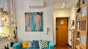 Un canapé de couleur turquoise agrémenté de coussins à motifs, un climatiseur fixé au mur, une décoration murale en macramé et une bibliothèque en bois dotée de portes en verre.