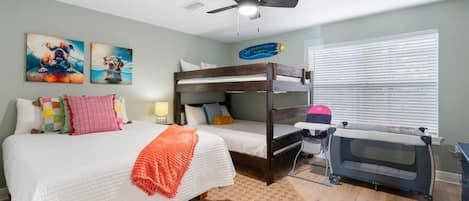Une chambre avec un lit superposé, un lit simple, un ventilateur de plafond, une fenêtre avec des stores et un panier pour animaux de compagnie.