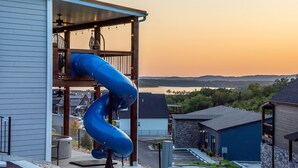 A residential area with a blue slide attached to a house, a covered porch, and a view of a lake at sunset. - Twisty slide, Lake view, Hot tub, Indoor pool, Fire pit | Skye Retreat (Branson)