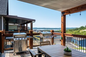 En overdækket terrasse med to grill i rustfrit stål, et træbord og udsigt til sø og bjerge.