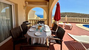 Terraço com mesa posta para refeição, cadeiras, um guarda-sol vermelho e vista para o mar.