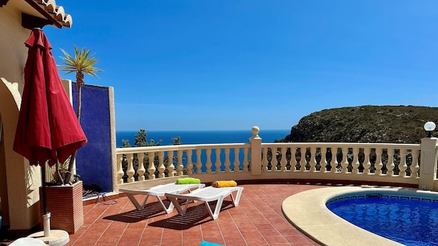Um terraço com piscina, cadeiras de descanso brancas e um guarda-sol vermelho.