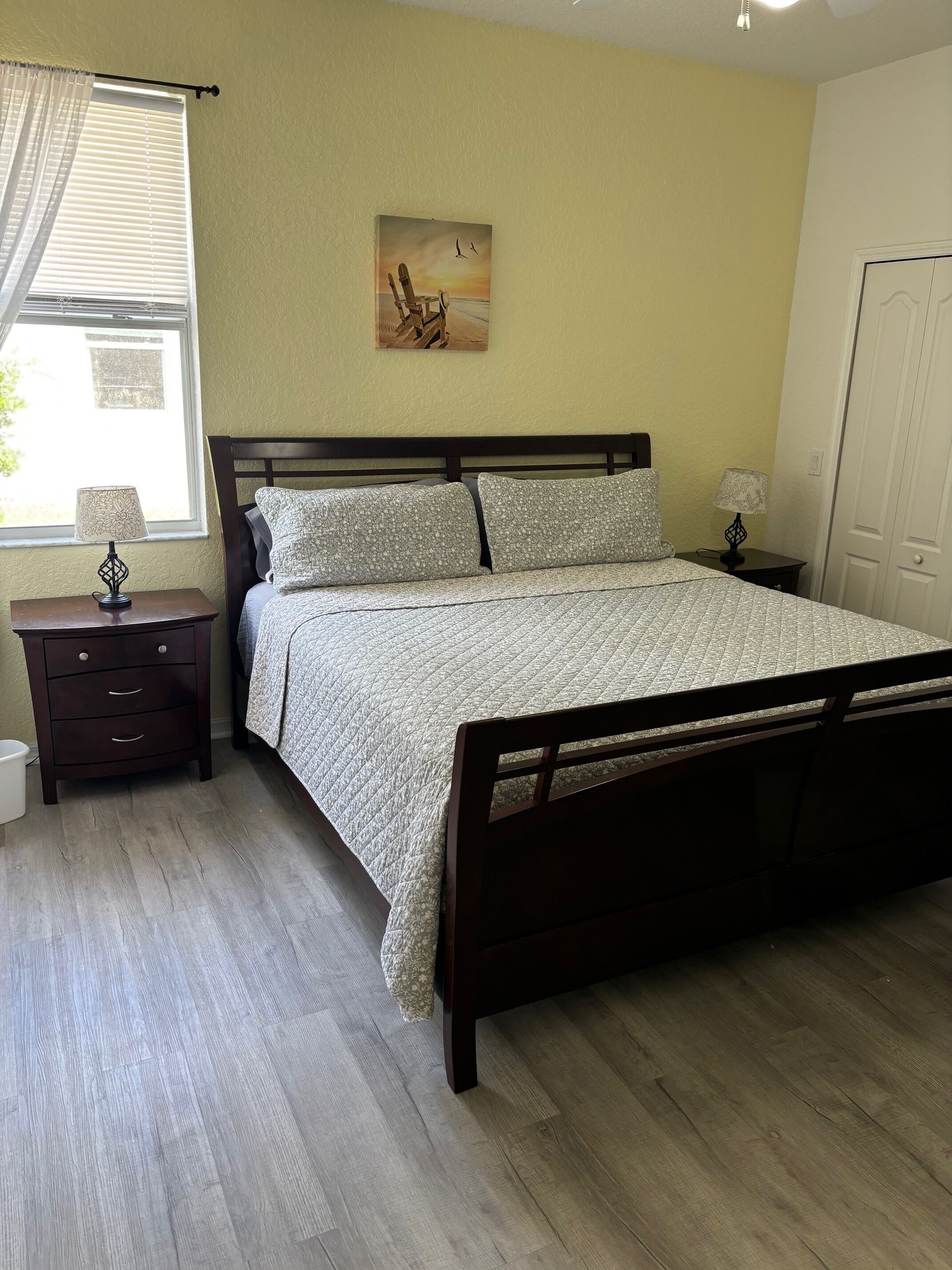 A bedroom with a bed, nightstands, a lamp, a painting, and a window with blinds.