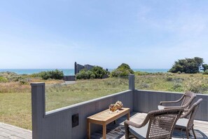 A deck with wicker chairs and a wooden table, overlooking a coastal landscape with a building in the distance.