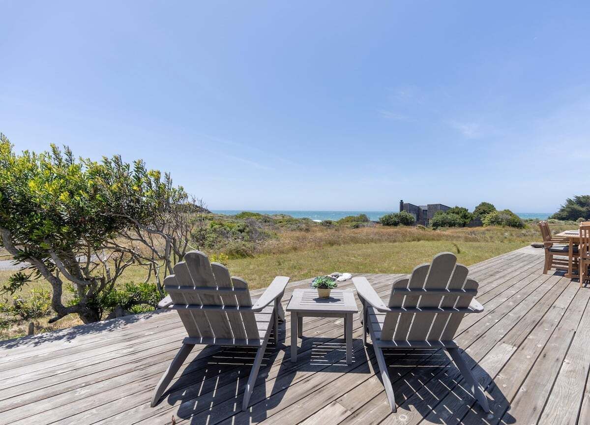 Una terraza de madera con dos sillas Adirondack y una pequeña mesa, con vistas a un paisaje costero bajo un cielo despejado.