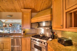 A kitchen with wooden cabinets, a stainless steel stove, a toaster, and a window with a view of trees. - Saltwater Serenity Lodge | Hot Tub & Views (Sea Ranch)