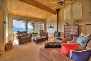 A cozy living room with a fireplace, comfortable seating, and a large window overlooking the ocean. - Saltwater Serenity Lodge | Hot Tub & Views (Sea Ranch)