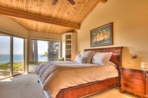 A bedroom with a large bed, a wooden ceiling, a painting, and a view of the ocean. - Saltwater Serenity Lodge | Hot Tub & Views (Sea Ranch)
