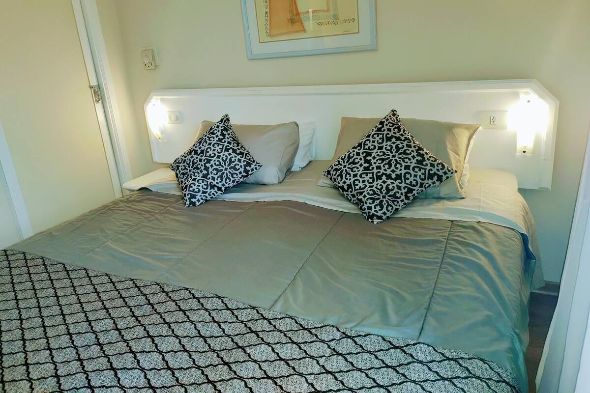 A neatly made bed with patterned pillows, a framed artwork above, and a simple bedside lamp.