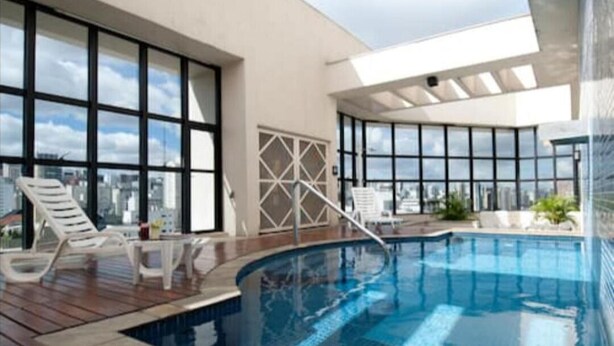 Una piscina interior moderna con paredes de vidrio transparente, una terraza de madera y sillones de descanso.