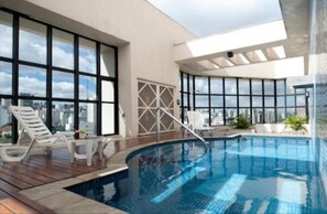 A modern indoor swimming pool with clear glass walls, a wooden deck, and lounge chairs. - Ninety Flat (São Paulo)
