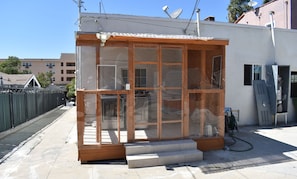 A small, enclosed structure with a wooden frame and glass panels, possibly a storage or utility room, with steps leading up to it and a hose on the ground.