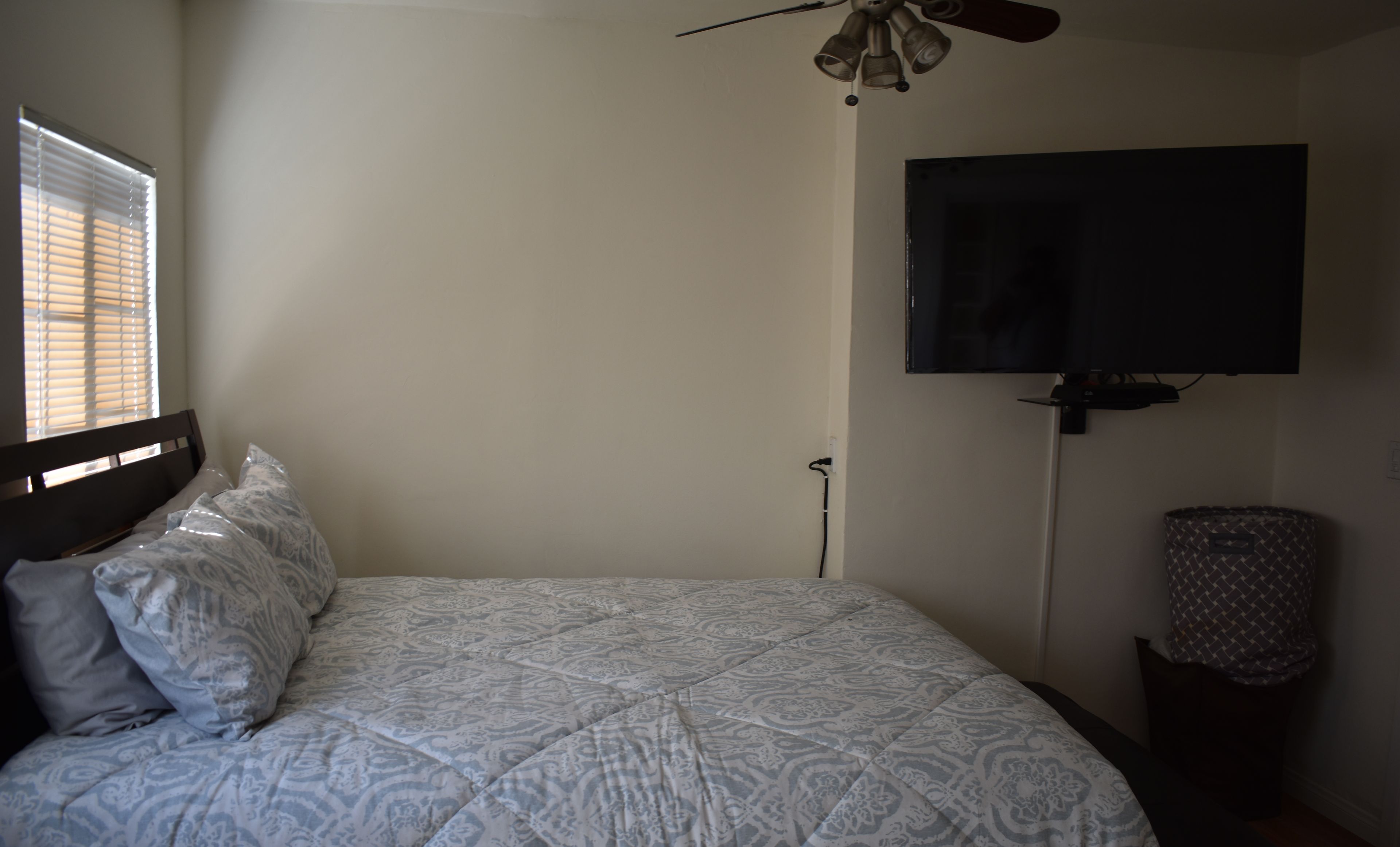 A bedroom with a bed, a ceiling fan, a TV mounted on the wall, and a window with blinds.