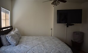 Ein Schlafzimmer mit einem Bett, einem Deckenventilator, einem an der Wand montierten Fernseher und einem Fenster mit Jalousien.