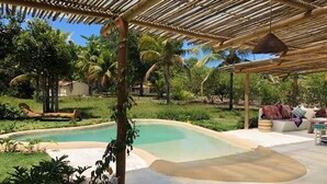 Ein Poolbereich mit einem Reetdach, umgeben von viel Grün und Palmen.
