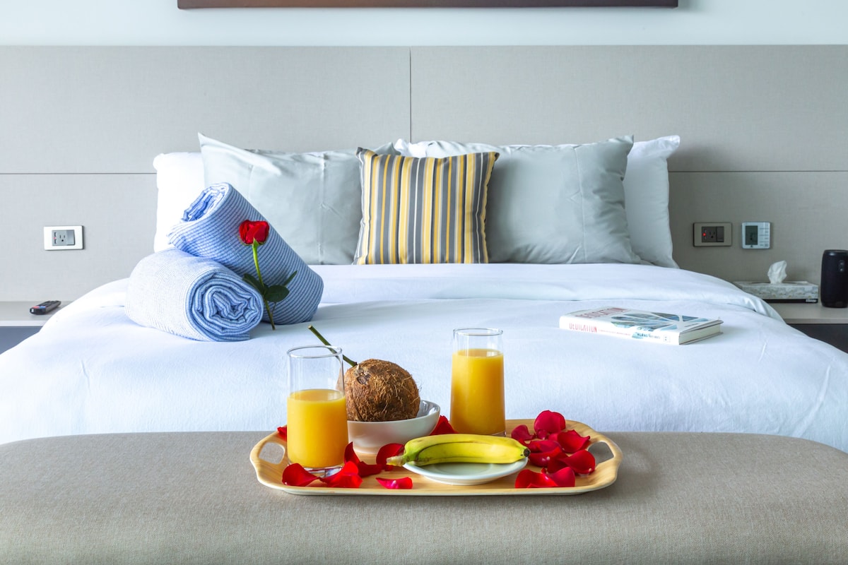 A neatly made bed with a pillow, a rolled towel, a book, a glass of orange juice, a bowl with a coconut, a banana, and a tray with rose petals.
