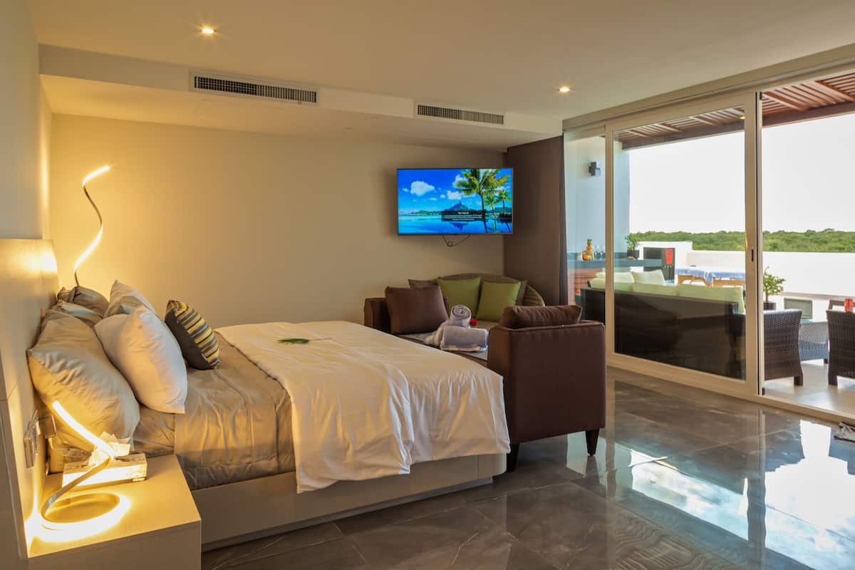 A modern hotel room with a bed, a sofa, a TV, and a balcony with a view of the sea.
