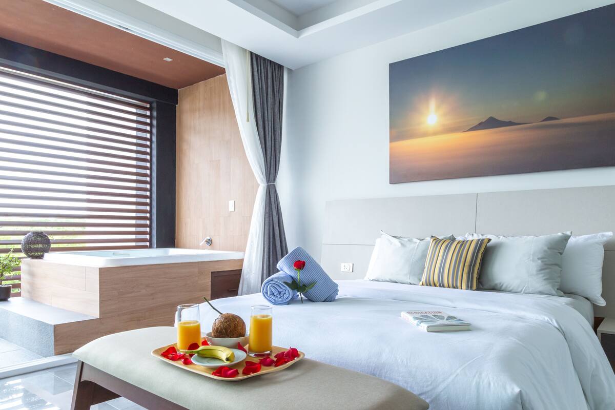 A hotel room with a bed, a breakfast tray with fruit and juice, a painting of a sunset, and a view of the outdoors through the window.