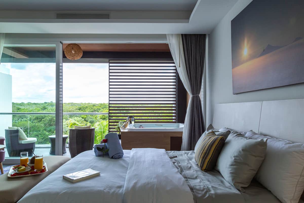 A modern hotel room with a large bed, a bedside table, a window with blinds, a view of greenery, and a painting on the wall.