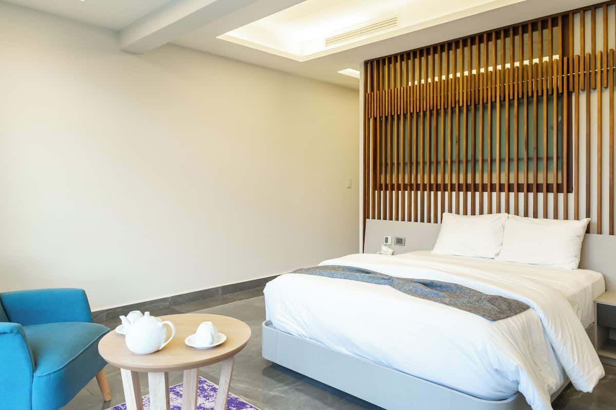 A modern hotel room with a bed, a blue armchair, a small round table with a teapot and cups, and a wooden slat wall design.