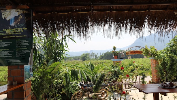 A rustic outdoor seating area with a thatched roof, a motorcycle, and a signboard for an adventure tour. - Phong Nha Friendly Home 2 (Phong Nha)