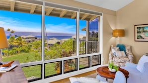 A room with large windows offering a view of a tropical landscape and ocean. There is a sofa, a table with a plant, and a lamp. - Molokai views, refreshed upgrades, relax comfort (Lahaina)