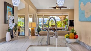 A modern kitchen with a marble countertop, stainless steel faucet, and a bowl of fruit. There is a ceiling fan and a map on the wall. - Molokai views, refreshed upgrades, relax comfort (Lahaina)