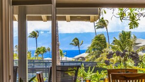 A table with a glass of wine and a menu, overlooking a tropical garden with palm trees and a view of the ocean. - Molokai views, refreshed upgrades, relax comfort (Lahaina)