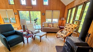 A cozy living room with wooden walls and large windows, featuring a sofa, chairs, a fireplace, and a telescope.