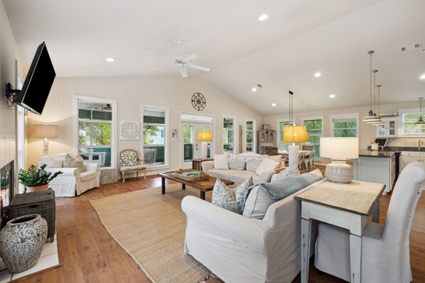 A spacious living room with a large sofa, a coffee table, and a dining area with a table and chairs. There is a kitchen area with cabinets and a window.