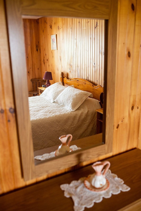 A wooden-framed mirror reflecting a bedroom with a bed, pillows, a nightstand, and a lamp.