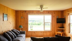 Una habitación con paneles de madera, un ventanal grande, un ventilador de techo, un televisor sobre un estante de madera y un sofá de color oscuro.