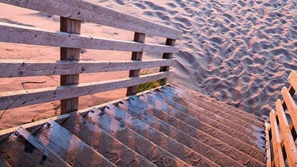 Una escalera de madera que baja hacia una playa arenosa con huellas, una barandilla de madera y un atardecer de fondo.