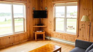 Habitación con paredes de madera, un televisor de pantalla plana montado, una pequeña mesa de madera, un sofá y ventanas amplias con vista a un área verde y edificios.