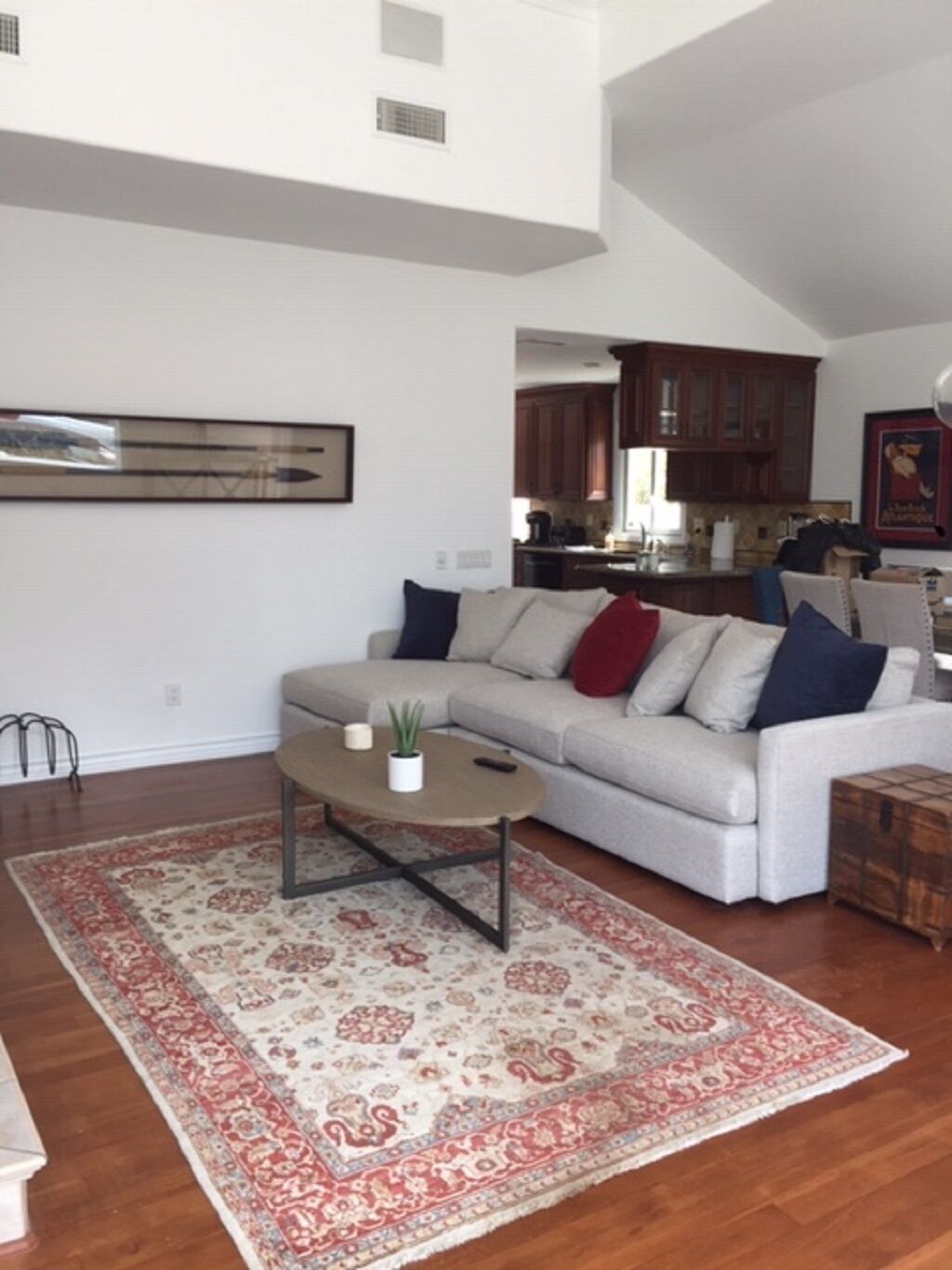 A spacious living room with a grey sofa, a coffee table, and a patterned rug. A kitchen is visible in the background with wooden cabinets.