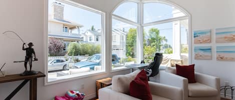 A modern living room with a large bay window, a white sofa, a red pillow, and a statue on a wooden table.