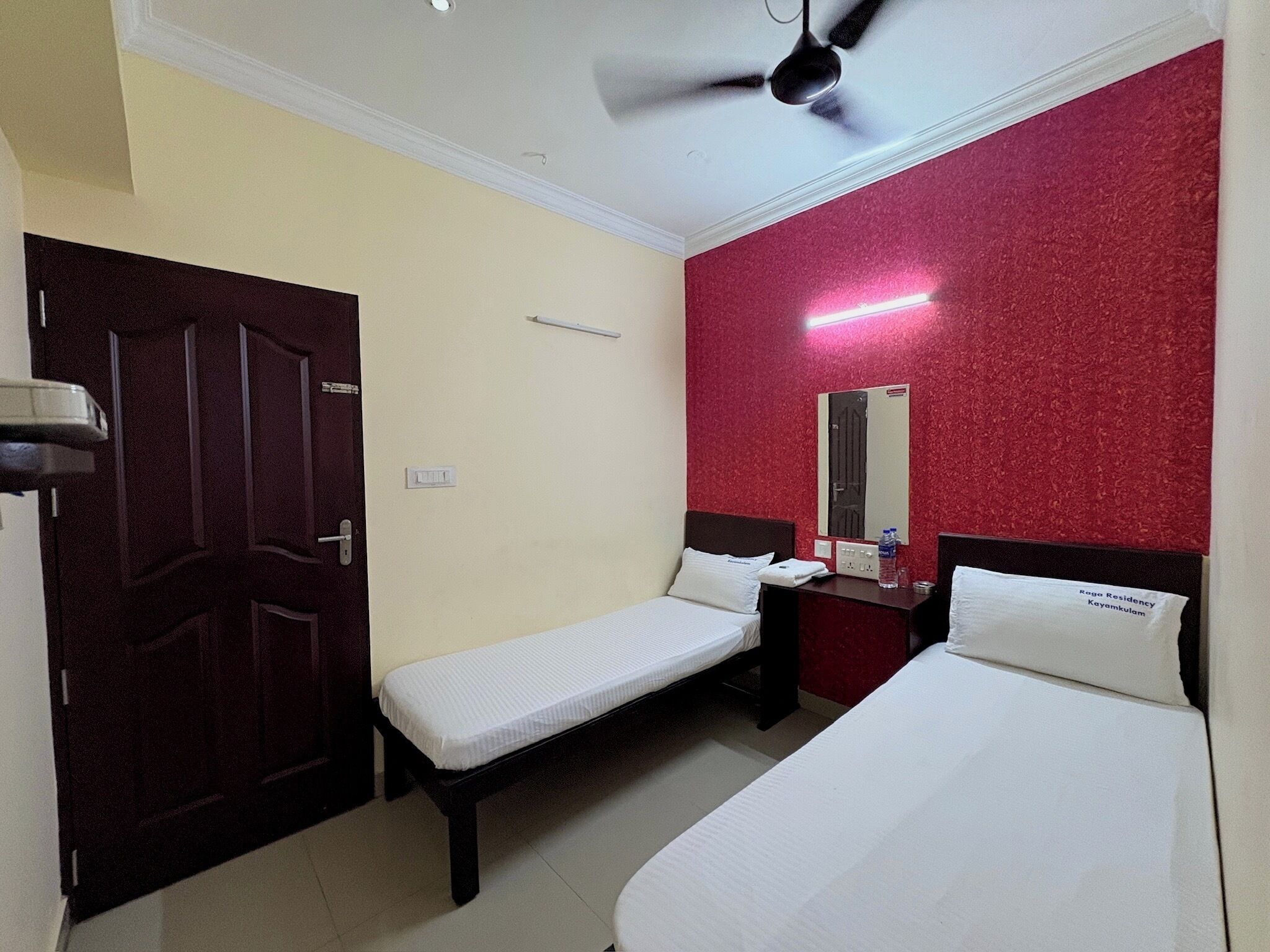 A hotel room with two single beds, a ceiling fan, a red accent wall, a mirror, and a nightstand with a phone and a bottle.