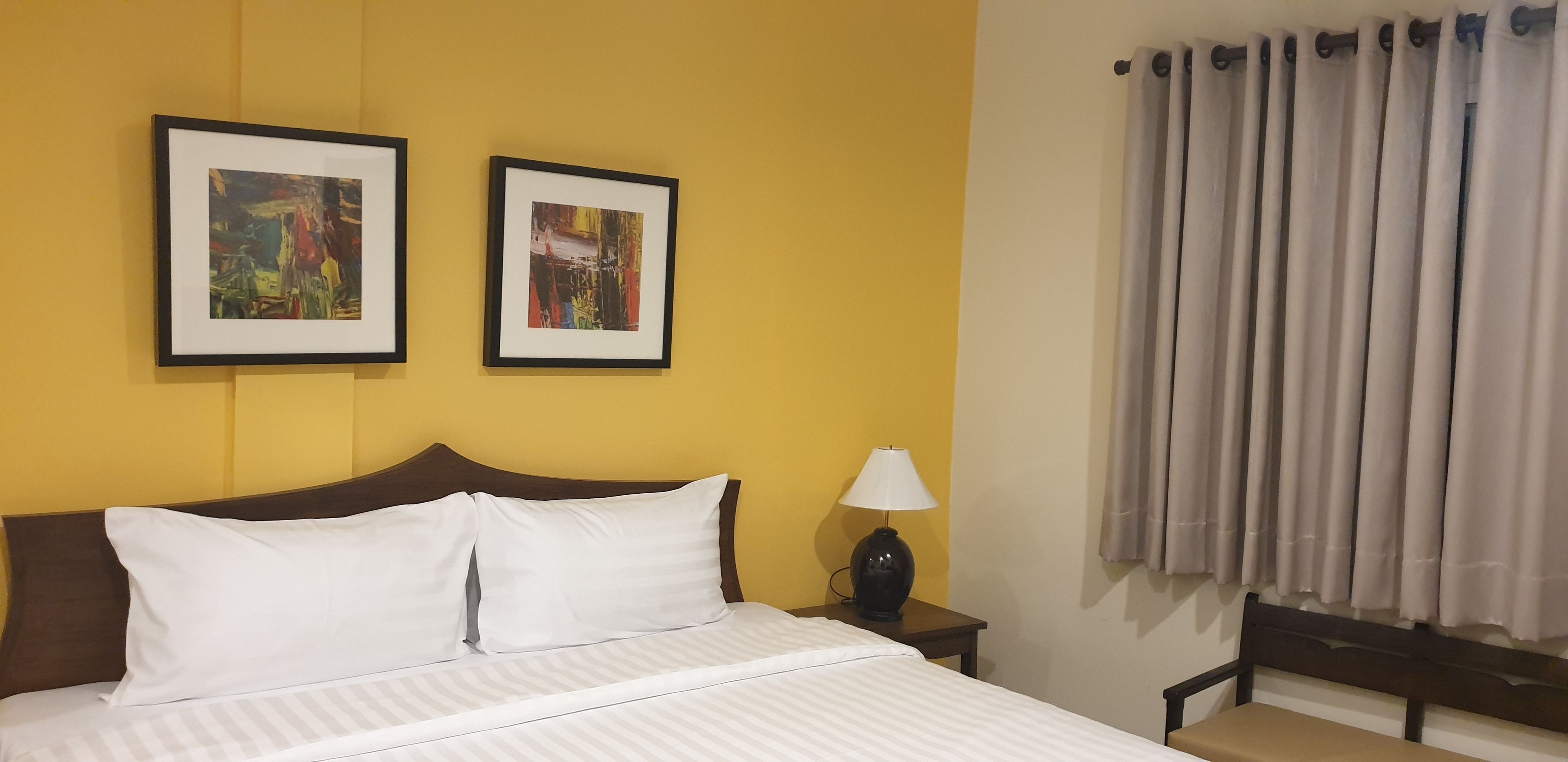 A hotel room with a bed, two framed abstract paintings on the wall, a nightstand with a lamp, and a window with curtains.