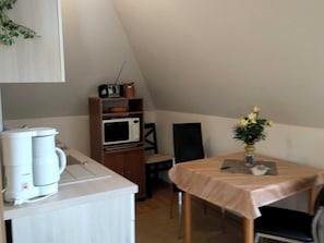 A small, cozy kitchen with a microwave, coffee maker, and a shelf with a radio and a pot. There's a table with a vase of flowers and a chair. - Morning sun - Apartments Meeresfrische (Kühlungsborn)
