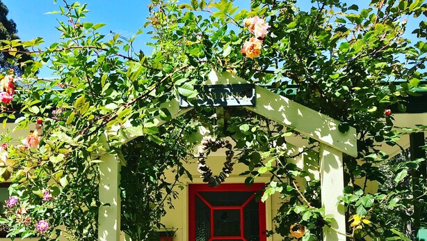 Una puerta roja con un adorno en forma de corazón, un letrero sobre la puerta y un arco de jardín con plantas trepadoras.