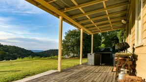 A covered outdoor area with a wooden deck, a grill, and a view of a green landscape.
