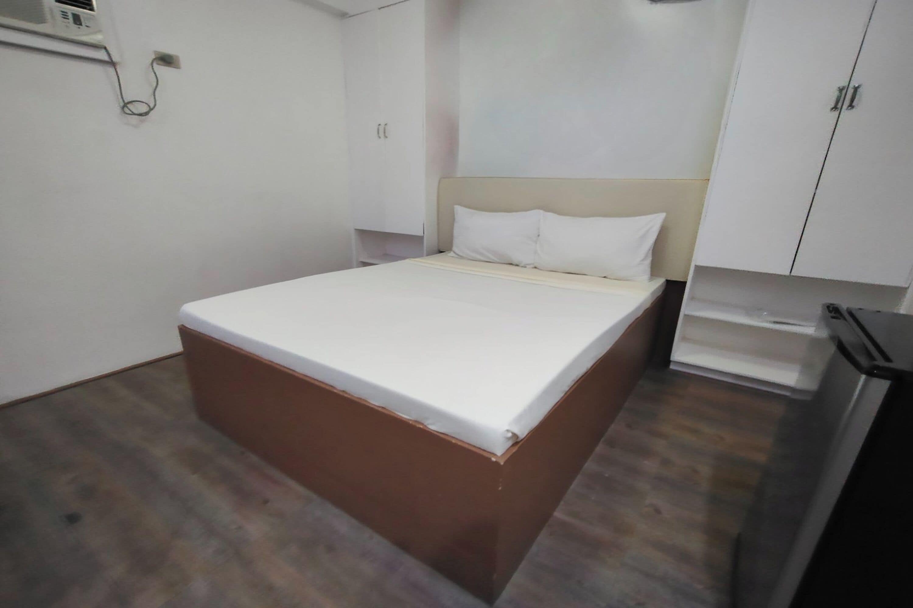 A single-bed room with a wooden floor, white walls, a beige headboard, and a white mattress.