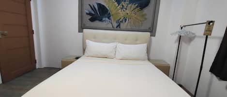 A bedroom with a bed, two white pillows, a beige headboard, a wall-mounted artwork, a nightstand, a bedside table, and a hanging lamp.