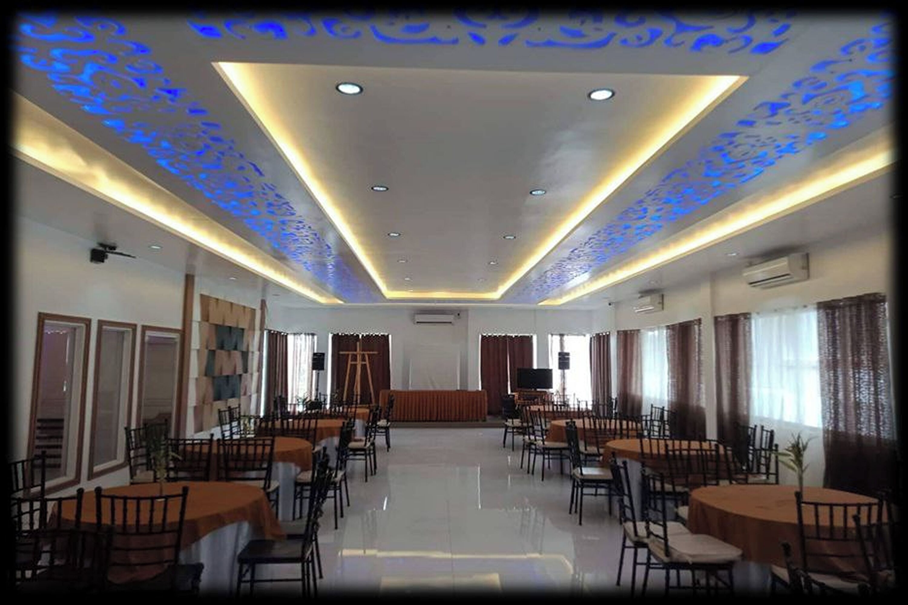 A spacious conference room with multiple tables and chairs, a stage, and a ceiling with blue light patterns.
